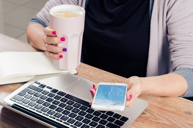 Photo image of a person at a laptop while looking at their phone and holding a big cup of coffee.