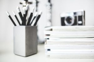 A photograph of a desk with a pencil holder, filled with black pencils, a stack of white paper with a camera on top of the stack.