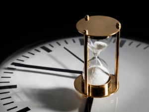 This is a photograph of an hourglass in a gold frame with white sand set on top of a large clock face.